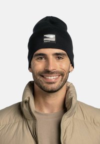 Black knit beanie with a round top and a rectangular patch featuring a mountain and sun design, worn by a smiling individual in a tan jacket.