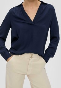 Marineblaue Bluse mit langen Ärmeln und einem eingeschnittenen Kragen. Weicher Stoff, leichter Glanz, lockere Passform. Kombiniert mit hellbeigen Hosen mit Knopfakzenten.