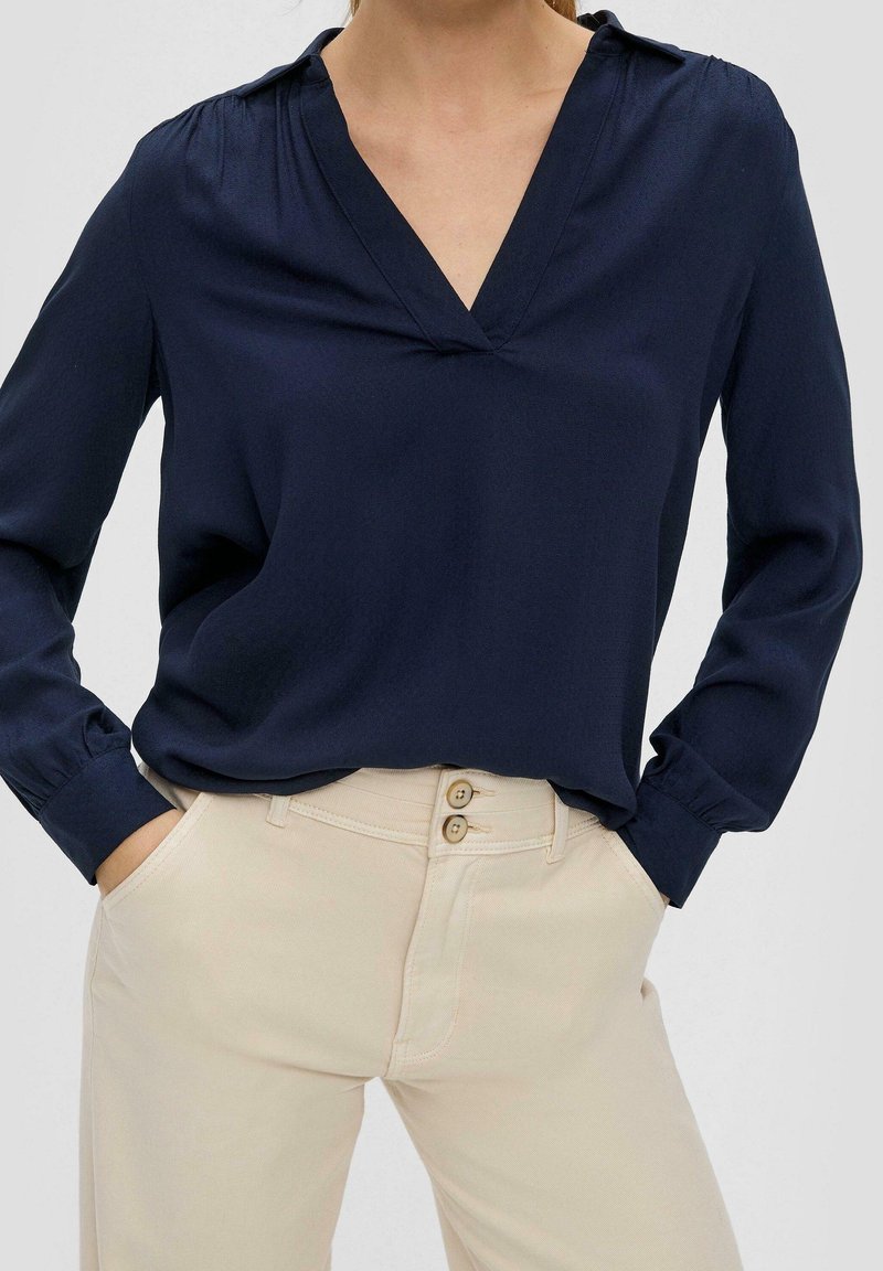 Marineblaue Bluse mit langen Ärmeln und einem eingeschnittenen Kragen. Weicher Stoff, leichter Glanz, lockere Passform. Kombiniert mit hellbeigen Hosen mit Knopfakzenten.