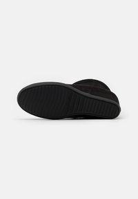 Botte en suède noir avec une semelle en caoutchouc texturée, présentant une pointe arrondie et un design minimaliste. La botte possède une doublure intérieure douce et moelleuse.