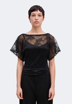 Personne portant un haut en dentelle noire avec des motifs floraux et des manches courtes transparentes, assorti à un pantalon noir, debout devant un fond uni.