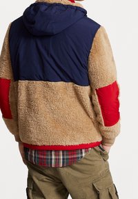 Giacca fuzzy color sabbia con cappuccio blu, maniche rosse e dettagli beige. Presenta una finitura testurizzata e una camicia a quadri a strati sottostante.