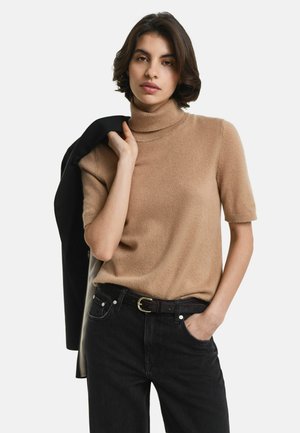 Frau mit kurzen dunklen Haaren, bekleidet mit einem beigen T-Shirt mit kurzen Ärmeln und Stehkragen, schwarzen Jeans mit Gürtel, die eine schwarze Jacke über ihrer Schulter hält.