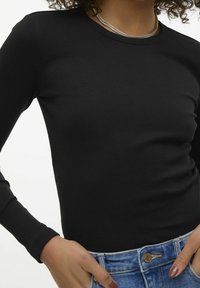 Eine Person trägt ein enges, schwarzes langärmliges geripptes Shirt und blaue Jeans mit den Händen, die nahe dem Bund ruhen, sowie eine silberne Kettenuch.