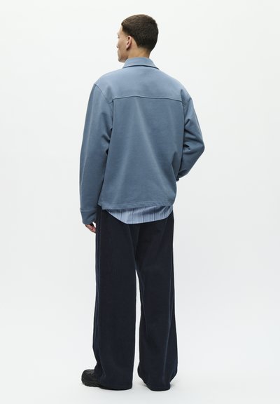 Man wearing light blue jacket over striped shirt and loose dark pants, standing facing away against plain white background.
