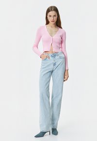 Cardigan corto rigato rosa con scollatura a V profonda e chiusura con bottoni, abbinato a jeans a vita alta blu chiaro e scarpe con punta blu.