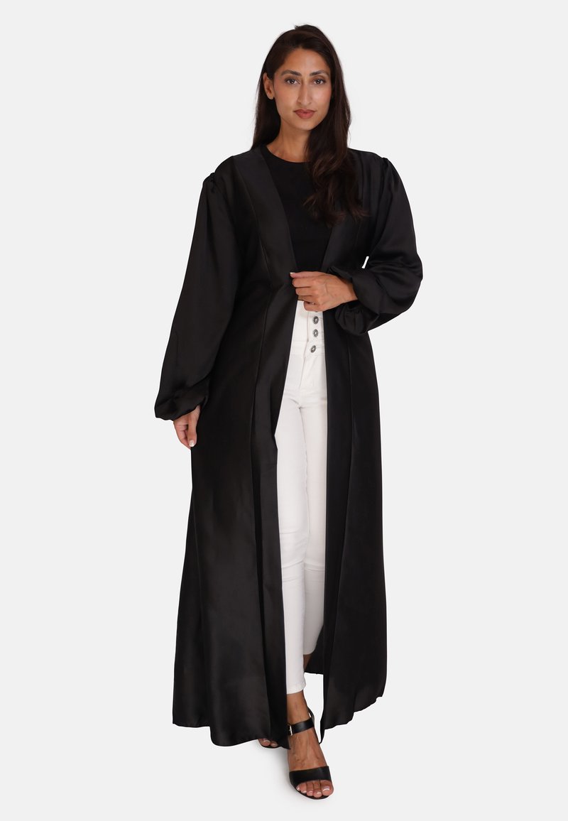 Sort satin lang frakke med pufærmer, V-hals design, åben front og en flydende silhuet, parret med hvide bukser og sorte sandaler.