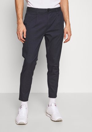 Man met zwarte slim-fit broek, witte sokken en witte sneakers, staand tegen een effen witte achtergrond.