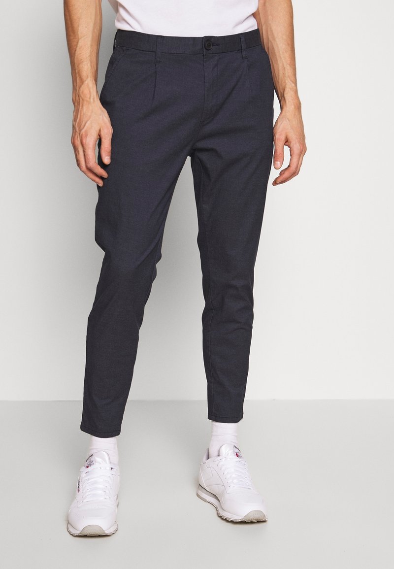 Homme portant un pantalon slim noir, des chaussettes blanches et des baskets blanches, debout devant un fond blanc uni.