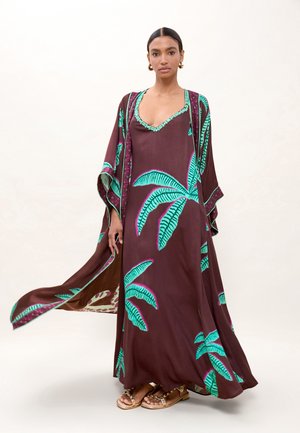 Mujer con un vestido largo marrón fluido con grandes estampados de hojas de palma verdes, combinado con sandalias doradas y pendientes de aro, posando sobre un fondo neutral.