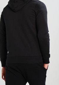 Sudadera de algodón negra con capucha ajustable, dobladillo y puños acanalados. Diseño simple, sin logotipos visibles ni detalles intrincados.