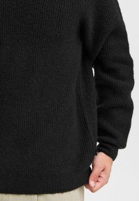 Black knitted sweater with ribbed texture, relaxed fit, and long sleeves. Features a side slit and a small brand tag near the hem.