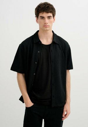 Joven con cabello rizado que lleva una camisa negra de manga corta acanalada sobre una camiseta negra, de pie frente a un fondo claro y liso.