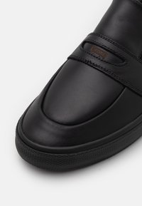 Svart lädersneaker med rund tå, slät yta, elastiska remmar och diskret logotyp på en brun etikett. Gummisula med texturerad yta.