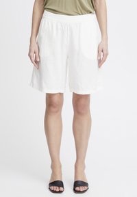 Shorts en lin blanc avec une taille élastique, offrant une coupe décontractée et une longueur mi-cuisse, assortis à des sandales à glissière noires.