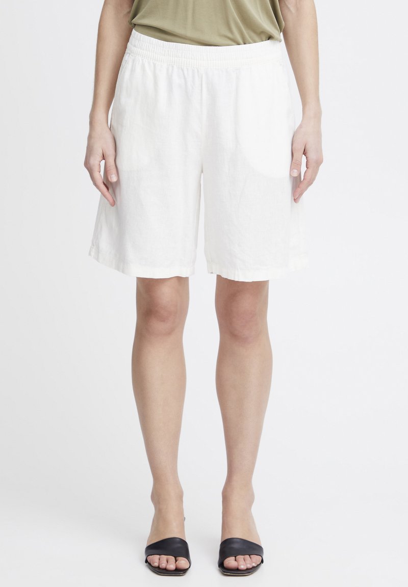 Shorts en lin blanc avec une taille élastique, offrant une coupe décontractée et une longueur mi-cuisse, assortis à des sandales à glissière noires.