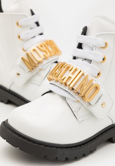 Valged patendinahast pahkluu saapad musta kummist tallaga, millel on kuldsed logo rihmad ja valged nöörid. Õmblemine on nähtav.