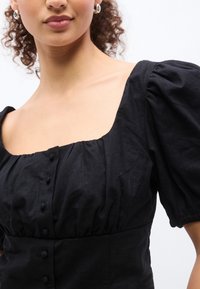 Femme portant une robe noire à encolure carrée avec des manches courtes bouffantes et des boutons recouverts de tissu sur le devant, visible des épaules à la taille.
