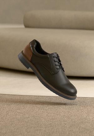 Chaussure habillée en cuir noir et marron à lacets flottant au-dessus d'un tapis beige avec un canapé beige en arrière-plan.