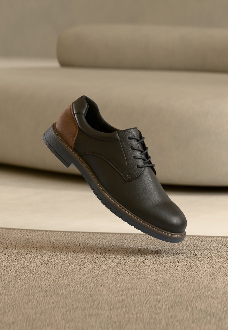 Chaussure habillée en cuir noir et marron à lacets flottant au-dessus d'un tapis beige avec un canapé beige en arrière-plan.