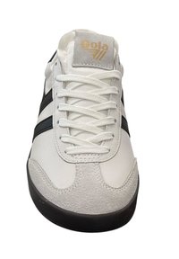 Weißer Sneaker mit schwarzen Streifen, aus Leder und Wildleder, runde Zehenpartie, sechs Ösen und einer strukturierten schwarzen Außensohle. Goldenes Logo auf der Zunge.