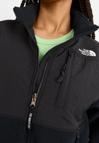 Svart fleecetröja med hög krage och fickor med dragkedja. Har en texturerad nedre del och en logotyp på axeln. Grön skjorta under.