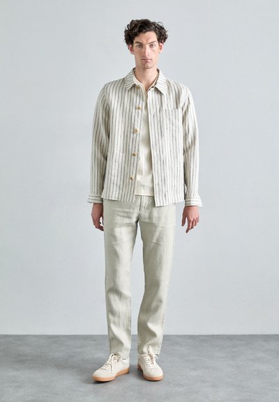Veste légère rayée en blanc cassé et noir, associée à une chemise neutre, un pantalon clair et des baskets beiges, mettant en avant une coupe décontractée.