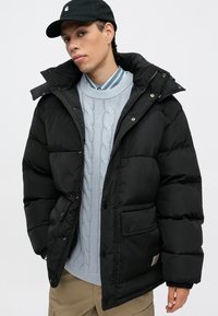 Fekete puffdzseki magas gallérral, elülső zsebekkel és patentos záródással. Alatta egy világoskék, kábelmintás pulóver látható.