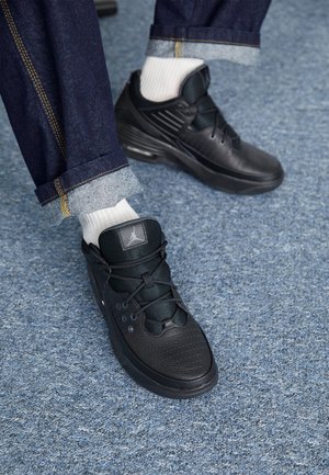 Personne portant un jean bleu foncé retroussé, des chaussettes blanches et des baskets Jordan noires debout sur un sol recouvert d'un tapis bleu.