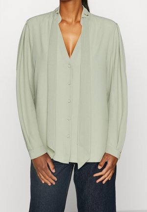 Kvinde iført en lysegrøn langærmet knappet bluse med slipsdetalje, kombineret med mørkeblå jeans, stående mod en ensfarvet baggrund.