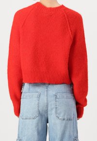 Suéter rojo de punto con un diseño recortado y dobladillo acanalado. Presenta mangas largas y un escote redondo. Combinado con jeans de tiro alto en un tono azul claro.