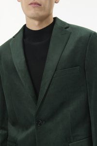 Žalias zomšinis švarkas su įpjautomis atlapomis, vieno mygtuko užsegimu ir krūtinės kišene. Dėvimas ant juodo golfelio. Lygus tekstūros paviršius.