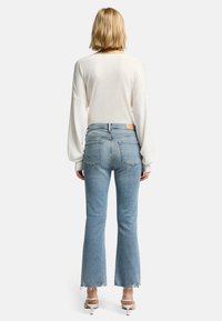 Jasno beżowa bluzka z długim rękawem w połączeniu z wyblakłymi, niebieskimi spodniami dżinsowymi typu dzwony, które mają surowe wykończenie i dwie tylne kieszenie. Spodnie kończą się powyżej kostki.