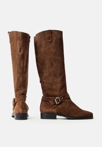 O pereche de cizme maro din suede, înalte, cu tocuri negre joase și curele cu catarame pe lateral, prezentate din lateral și din spate, pe un fundal alb.