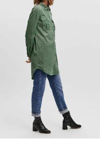 Vestito camicia in velluto a coste verde con due tasche sul petto, orlo arrotondato, abbinato a jeans blu arrotolati e stivaletti neri con tacco.