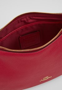 Sac en cuir rouge à surface texturée, avec fermeture éclair sur le dessus, ferrures dorées et doublure intérieure. Présente une étiquette de marque embossée à l'intérieur.