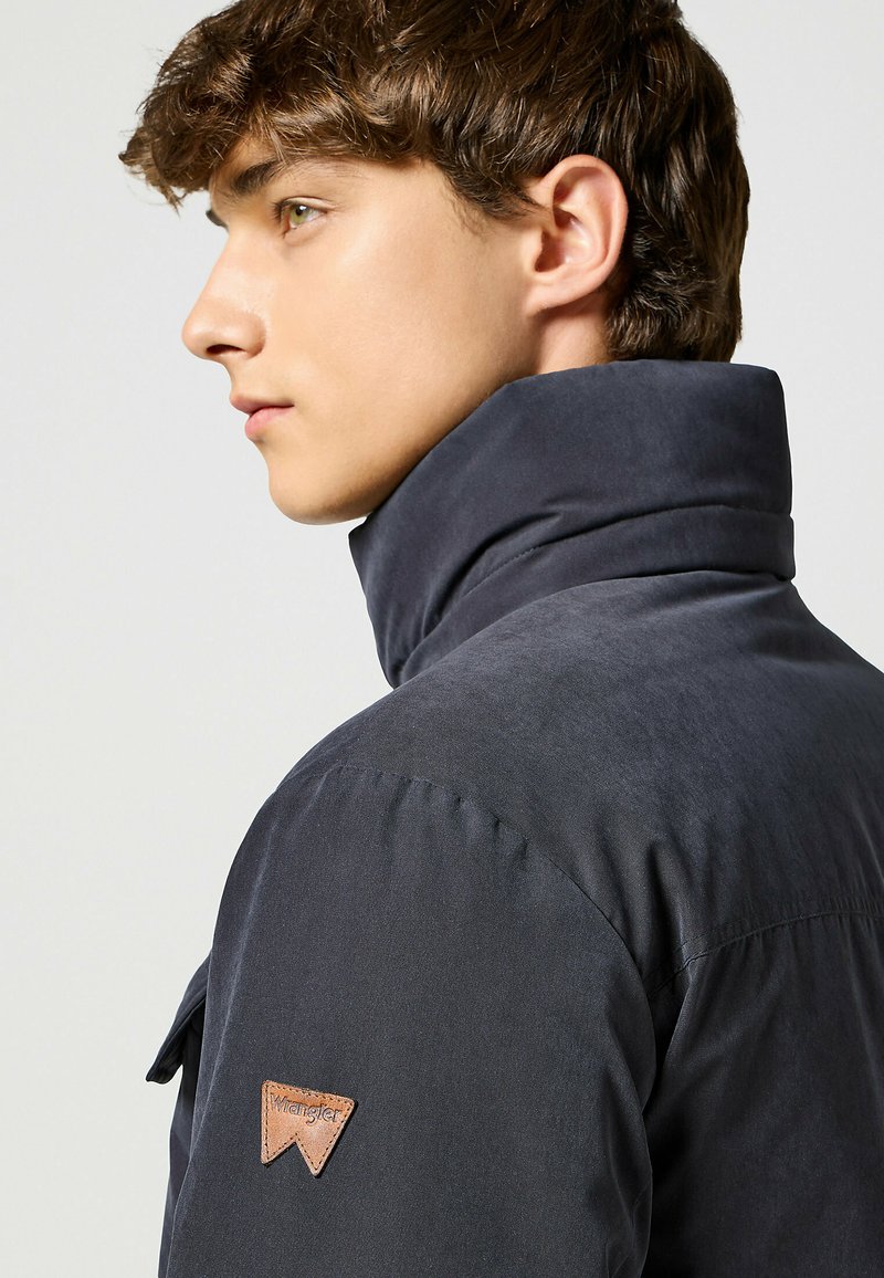 Giacca grigia scura con colletto alto, texture morbida e un patch in pelle sul spalla. Vestibilità su misura mostrata dalla vista laterale.