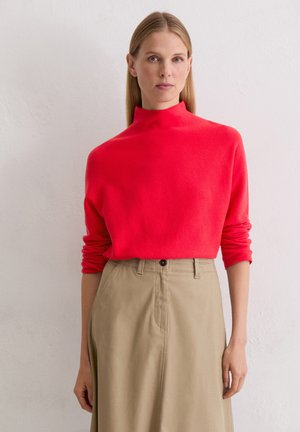 Red mock-neck sweater with rolled sleeves, paired with beige high-waisted skirt featuring a button closure and angled pockets.