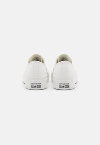 Fehér vászon, alacsony szárú sportcipő kerek orral, texturált felülettel és kontrasztos fekete "Converse All Star" logóval a saroknál.