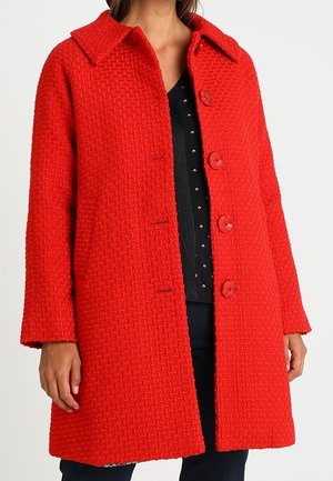 Woman wearing a textured bright red knee-length buttoned coat with a wide collar over a black top with small white and red patterns.