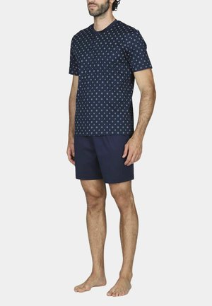 Uomo in piedi a piedi nudi che indossa una camicia a maniche corte fantasia blu navy e pantaloncini blu navy tinta unita su uno sfondo semplice.