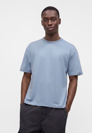 Homme debout devant un fond uni, portant un t-shirt bleu clair à manches courtes et un pantalon foncé, les mains dans les poches, de face.