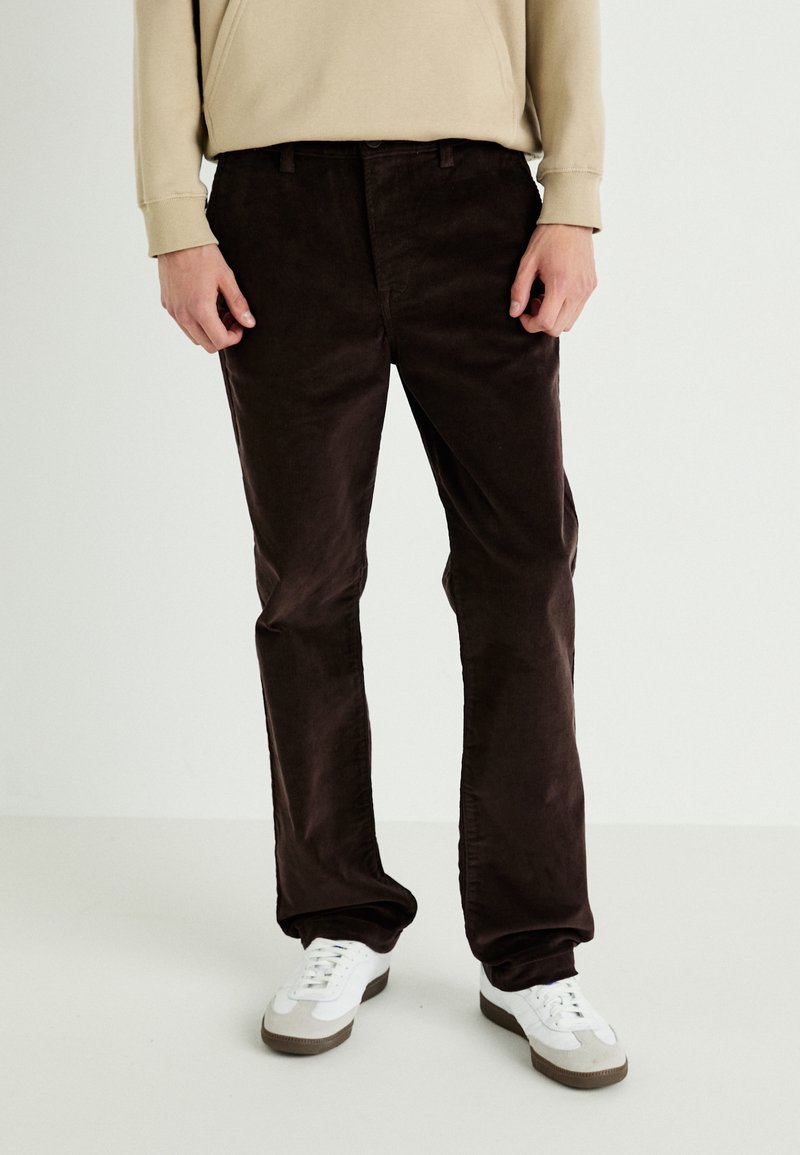 Bruine ribfluwelen broek met een rechte snit, met een textuur van ribbels, gecombineerd met witte sneakers en een beige hoodie.
