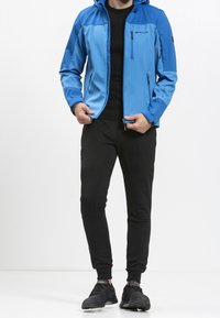 Modrá lehká bunda s kapucí; má kapsy na zip. Kombinováno s černými tepláky a černými sportovními botami.