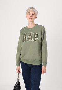 Felpa verde oliva con logo "GAP" in lettere scure. Presenta maniche lunghe e scollatura a coste, abbinata a jeans blu scuro e una borsa nera.