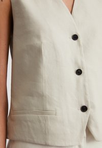 Helle beiger ärmelloser Weste mit V-Ausschnitt, versehen mit schwarzen Knöpfen und einer Seiten Tasche. Hergestellt aus einem strukturierten Stoff.