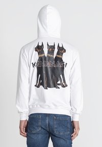 Biela mikina s kapucňou, na zadnej strane s veľkým grafickým znázornením troch psov plemena Doberman. Psy majú na sebe obojky a pod nimi je text "KAVANAGI".