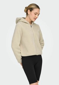 Beige fleece-hettejakke med glidelås, teksturert overflate og hette, kombinert med svarte ribbestrikkede shorts. Enkel design, uten synlig metallutstyr.