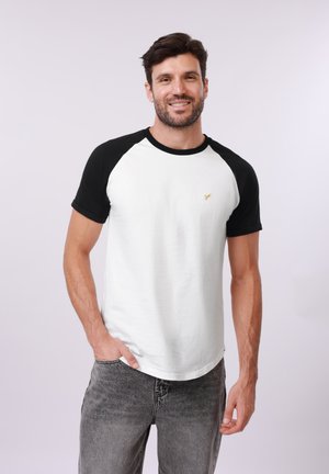 Lachende man staand met één hand in de zak, draagt een wit T-shirt met zwarte mouwen en grijze jeans, tegen een effen lichte achtergrond.