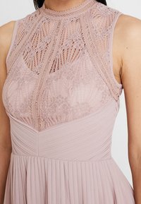 Roze jurk met een getextureerde kant bodice met ingewikkelde patronen, hoge halslijn en geplooide rok, gemaakt van lichte stof.